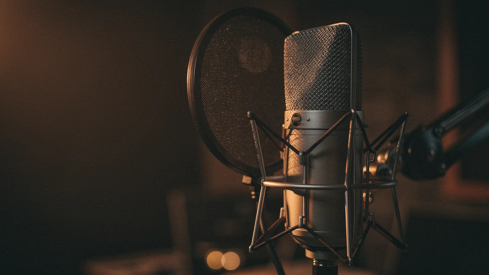 Vintage large-diaphragm condenser microphone in a dark studio with a black mesh pop filter in front, warm amber rim light catching the chrome grille of the microphone, an acoustic guitar barely visible in the deep bokeh behind