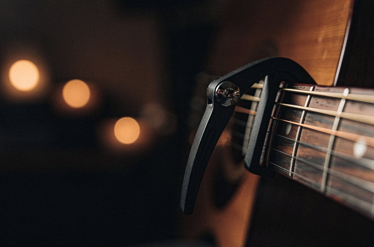 Black metal capo clamped across the second fret of an acoustic guitar neck, the strings pressed firmly behind the capo bar, warm amber side-light catching the chrome screw of the capo and the bronze of the strings, deep umber bokeh behind the neck