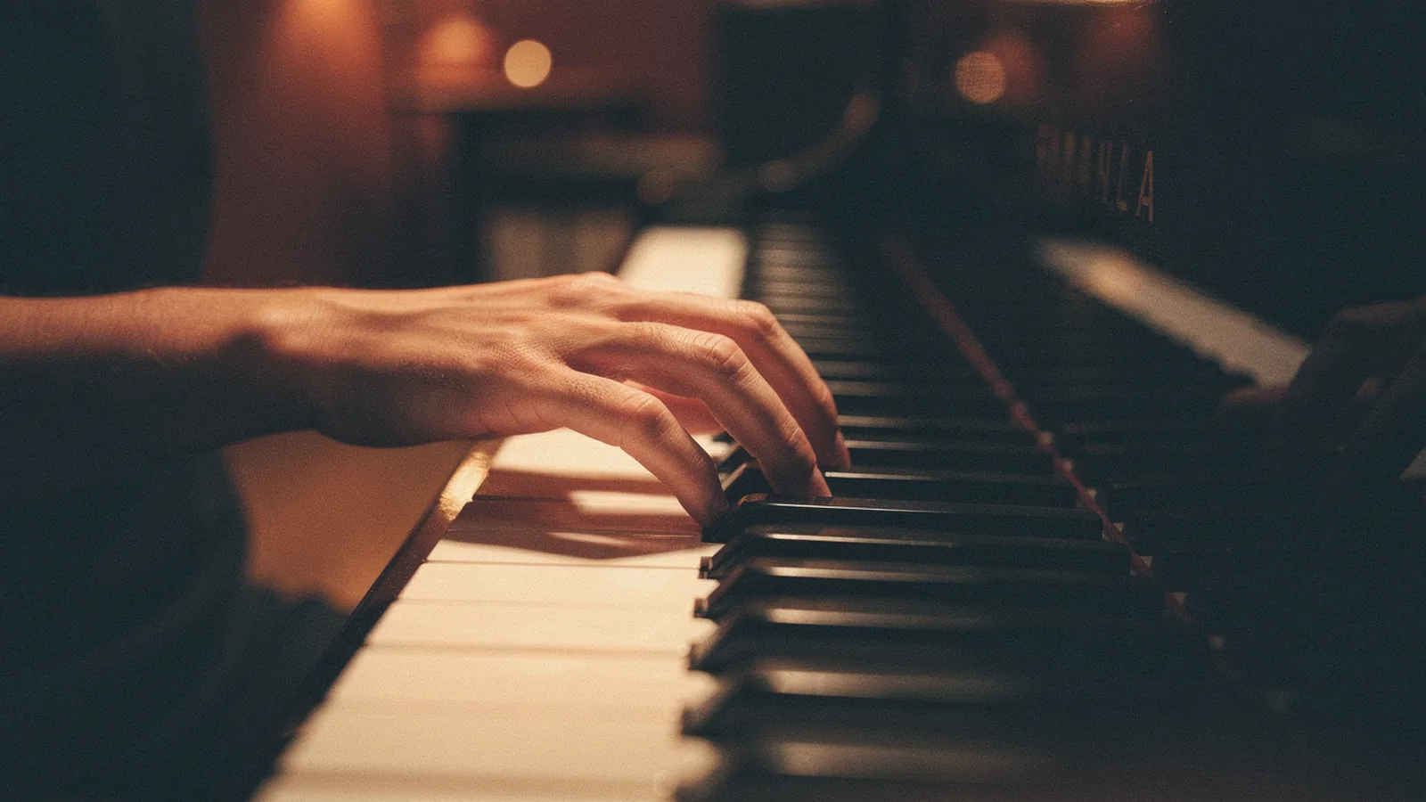 Hand pressing a chord on a piano keyboard, warm amber side-light from the left catching the contrast between ivory and ebony keys, the rest of the keyboard fading into deep bokeh and shadow