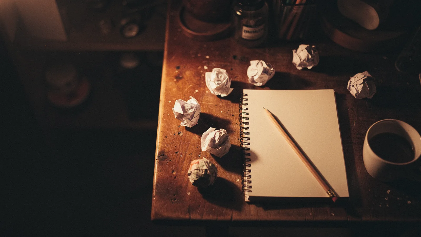 Open notebook with a completely blank page, a single sharpened pencil resting diagonally on the page, several crumpled paper balls scattered around it on a wooden desk, a half-empty coffee cup at the edge, dim warm amber lamp light fading into deep shadow