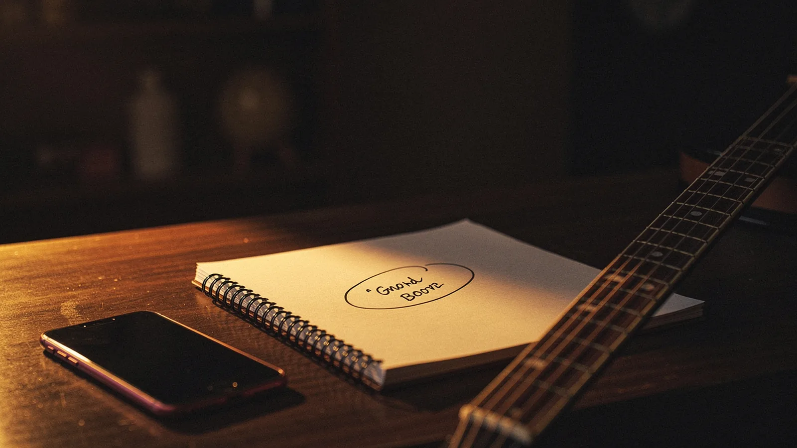 Open spiral notebook on a wooden desk with handwritten lyric lines and a circled title phrase, a smartphone propped face-up on the left, the neck and headstock of an acoustic guitar entering the right edge of the frame with strings and frets clearly visible, warm amber light catching all three songwriting seeds at once