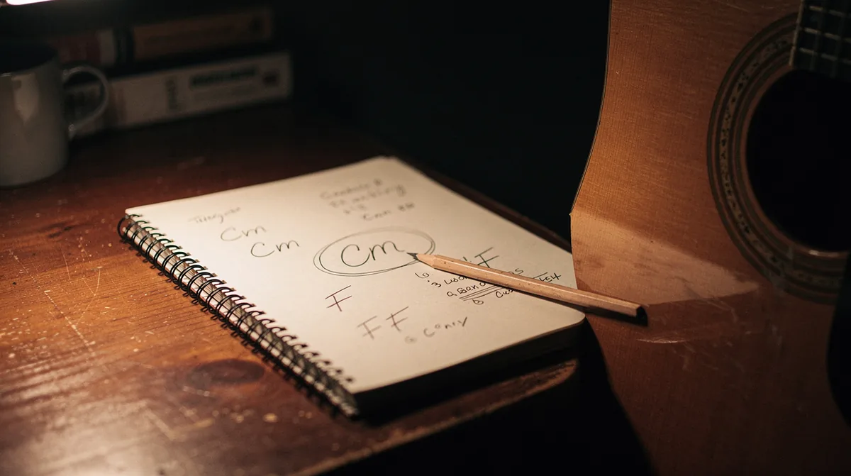 Open handwritten notebook on a wooden desk with chord names sketched in pencil including Cm and F written and circled, the body and soundhole of an acoustic guitar entering the right side of the frame, a pencil resting at an angle on the page, warm amber desk-lamp light fading into deep shadow on the left, intimate songwriter workspace