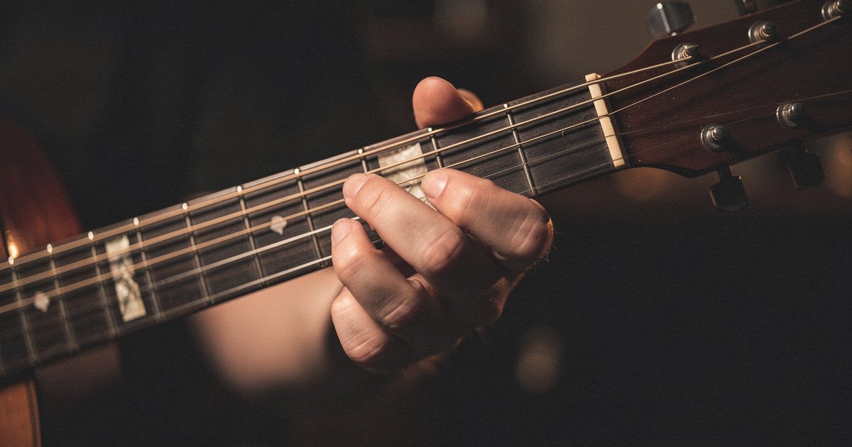 A hand pressing a chord shape on an acoustic guitar fretboard in warm amber light, the kind of voicing a songwriter reaches for when writing in a musical mode.
